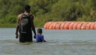 Migrantes caminan junto a grandes boyas que se utilizan como barrera fronteriza flotante en el río Bravo, el 1 de agosto de 2023, en Eagle Pass, Texas