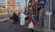 Una mujer que lleva una bolsa de compras junto a un trabajador de prevención de epidemias con un traje protector mientras continúan los brotes de COVID-19 en Beijing, China.