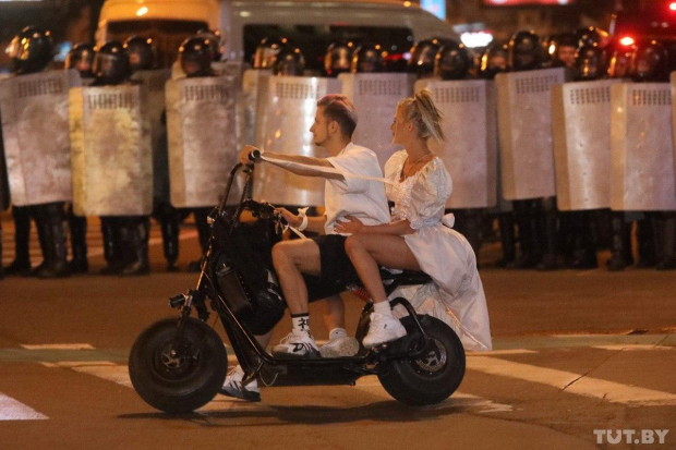 Una chica con vestido de novia pasa en una motoneta con su novio frente a un bloque de policías durante una manifestación en Minsk