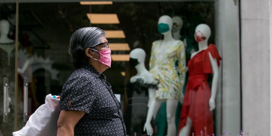 Una mujer que porta cubrebocas camina frente a unos aparadores donde también los maniquís usan mascarilla.