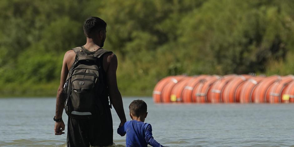 Migrantes caminan junto a grandes boyas que se utilizan como barrera fronteriza flotante en el río Bravo, el 1 de agosto de 2023, en Eagle Pass, Texas