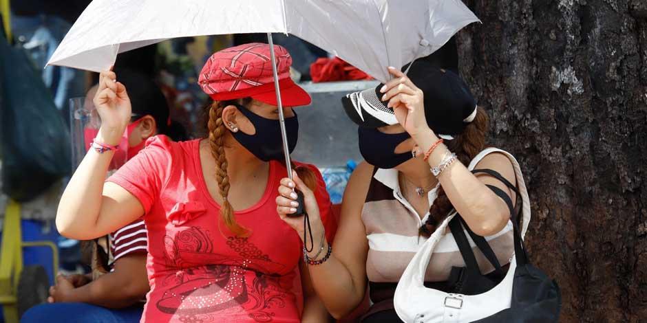 Dos mujeres se protegen del sol con un paraguas, y del COVID-19 con cubrebocas