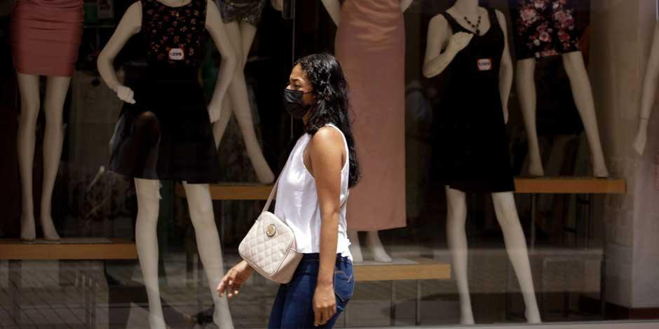 Una mujer que utiliza cubrebocas para protegerse de contagio por COVID-19 camina frente a un exhibidor de ropa