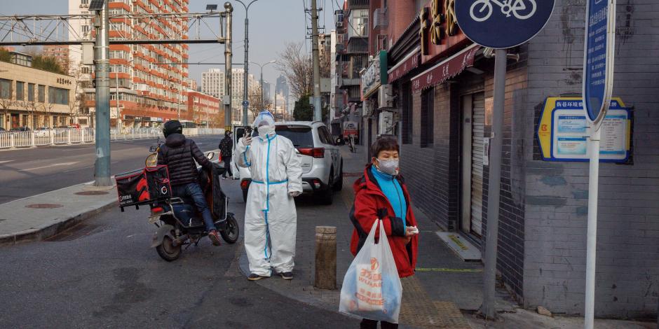 Una mujer que lleva una bolsa de compras junto a un trabajador de prevención de epidemias con un traje protector mientras continúan los brotes de COVID-19 en Beijing, China.