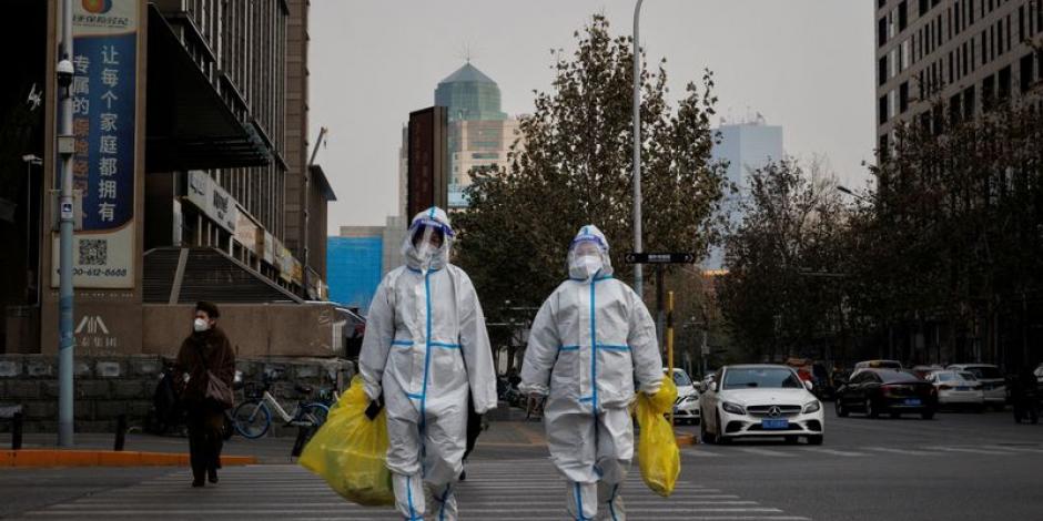 Trabajadores de prevención con trajes protectores cruzan una calle mientras continúan los brotes de la enfermedad por COVID-19.