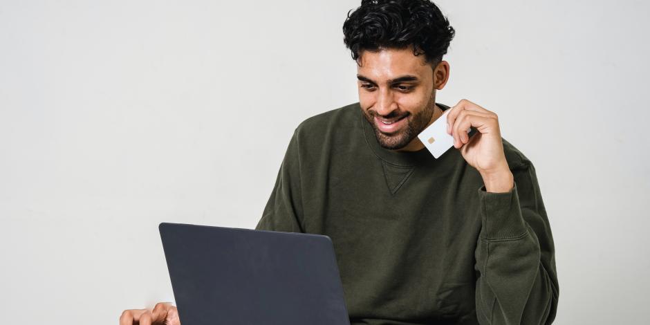 En la imagen ilustrativa, un hombre frente a una computadora sostiene una tarjeta en la mano para comprar en línea.