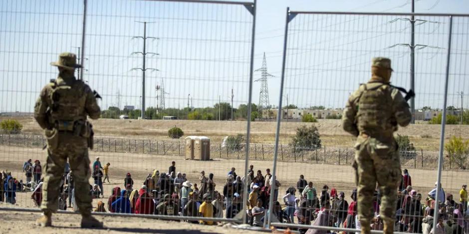 Migrantes esperan junto a una valla fronteriza mientras son vigilados por miembros de la Guardia Nacional de Texas, para ingresar a El Paso, Texas, el miércoles 10 de mayo de 2023.