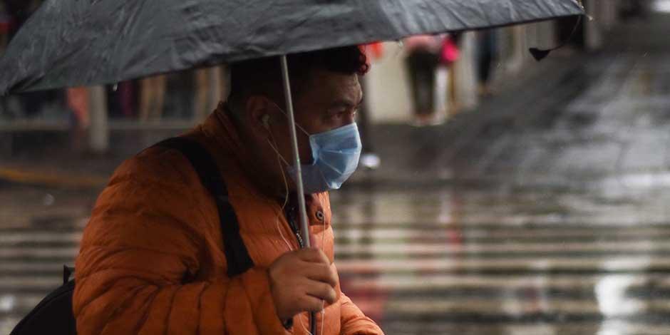 Una persona con cubrebocas camina portando un paraguas para protegerse de la lluvia
