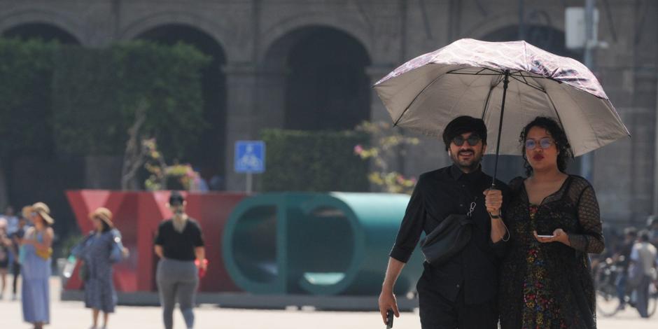 Una pareja camina por el Zócalo utilizando una sombrilla para cubrirse del sol.